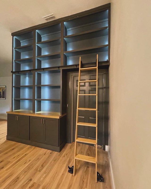 Dark green floor-to-ceiling built-in bookshelves with LED backlighting and a wooden rolling library ladder