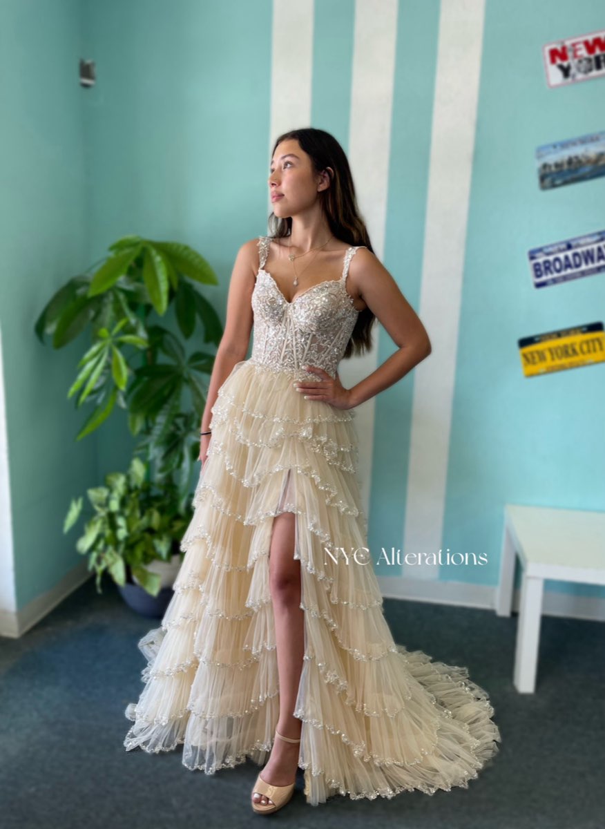 A client wearing a beaded, tiered champagne gown altered by NYC Alterations, standing in the shop's teal fitting room