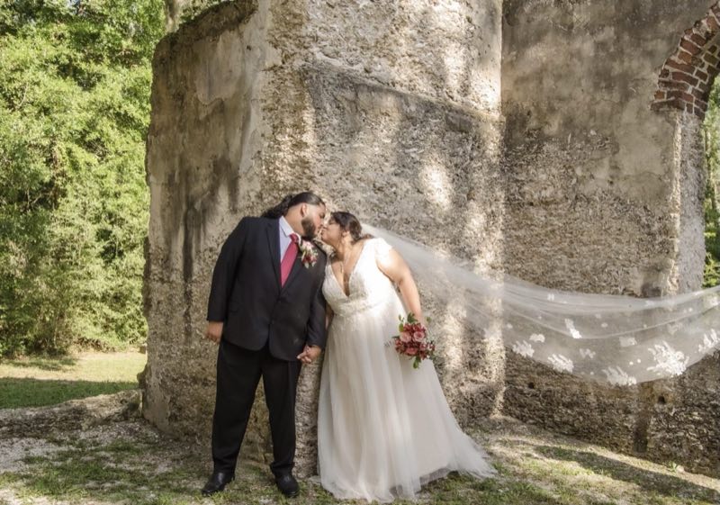 Newlywed couple kissing outdoors, the bride in a gown and long veil altered by NYC Alterations