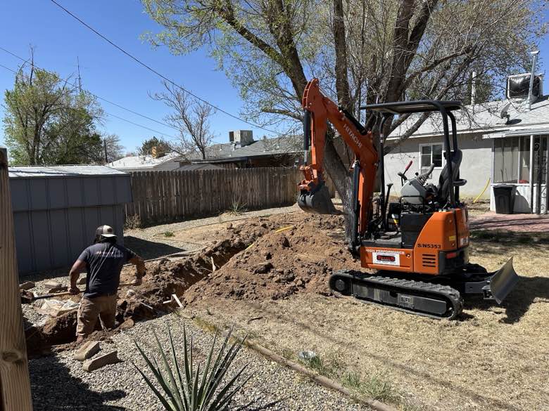 A worker and a mini-excavator digging a trench in a backyard for a sewer line replacement.